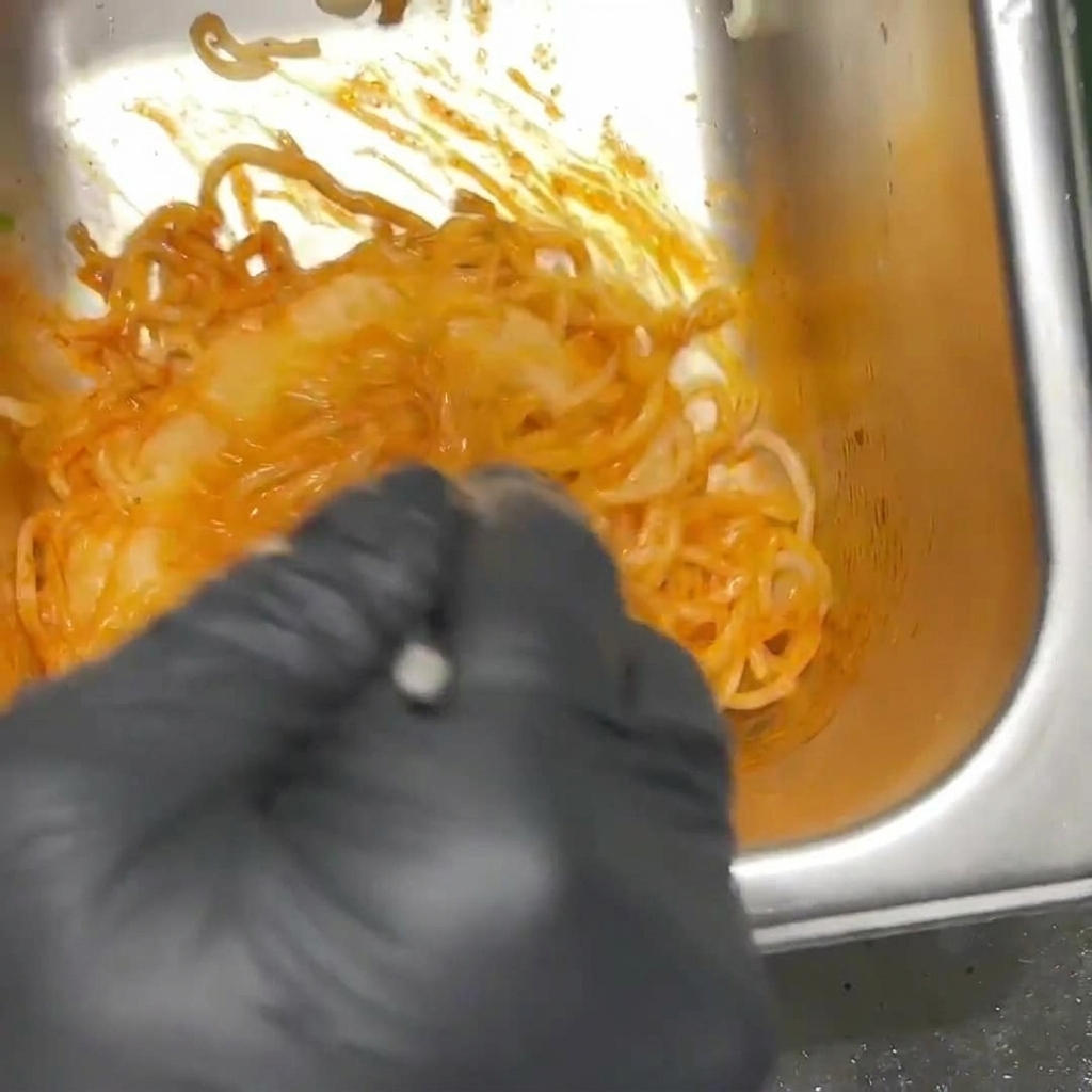 검은색 장갑을 낀 조리사가 스텐 볼 안에서 붉은 불닭 소스에 라면 면발을 골고루 버무려 면이 붉게 물든 모습.