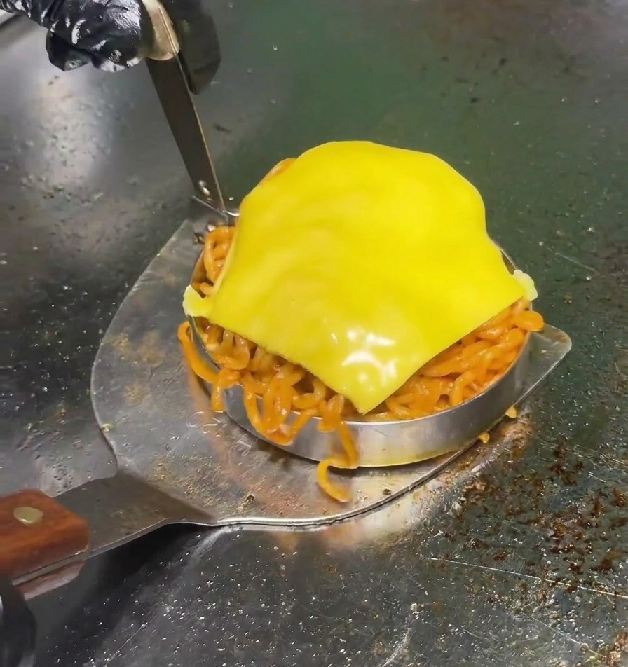 전문가용 그릴 위에서 노란 치즈가 녹아내린 매콤한 라면 패티를 금속 뒤집개로 몰드째 조심스럽게 들어 올리는 모습.