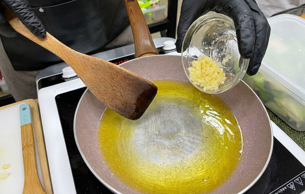 검은 장갑을 낀 손이 녹은 노란 버터와 오일이 담긴 팬에 작은 볼에 담긴 다진 마늘을 붓는 모습