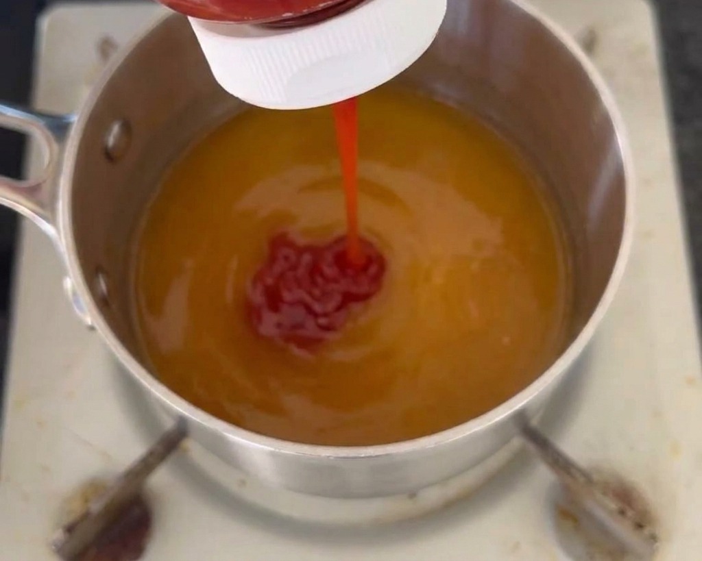 노란색 액체 소스 베이스가 담긴 냄비에 전용 용기에서 붉은 케첩을 짜서 넣는 모습.