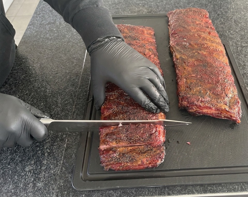 검은색 니트릴 장갑을 낀 사람이 검은색 도마 위에서 잘 익은 짙은 붉은색 훈제 폭립을 커다란 셰프 나이프로 뼈 사이사이를 썰어내는 모습.