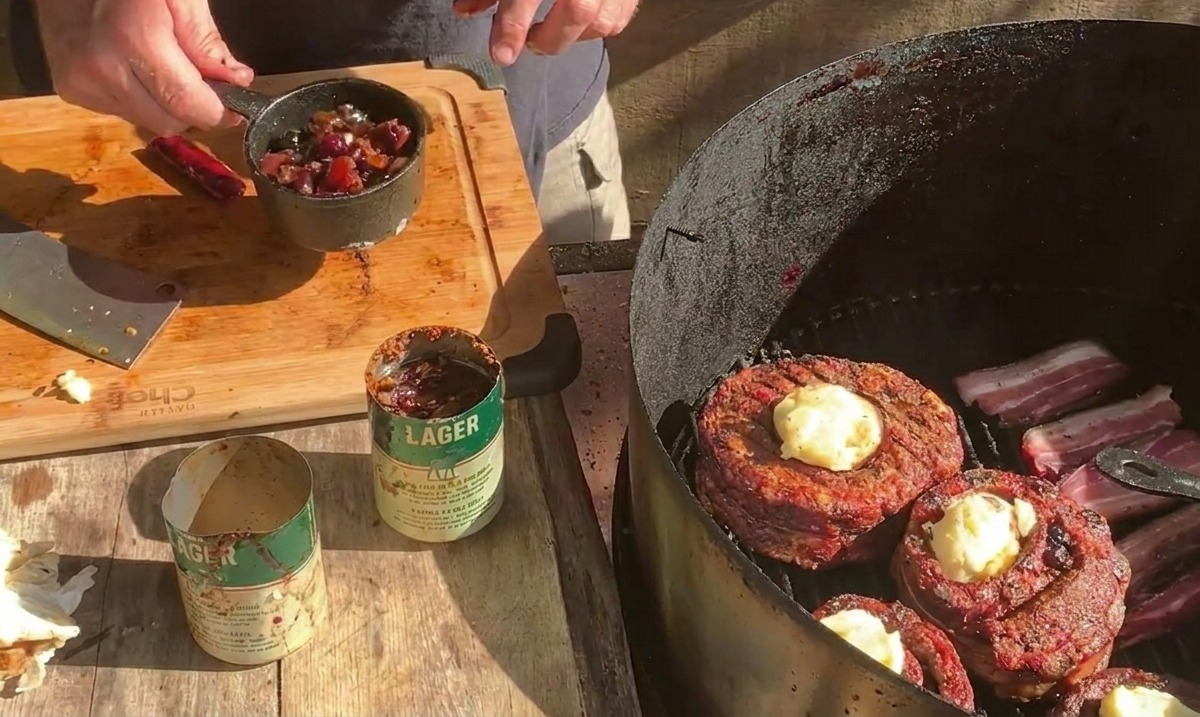 치즈가 뿌려지고 있는 베이컨을 감싼 립 컵 그릴 위로 블루베리 소스가 담긴 작은 금속 냄비를 들고 있는 모습.