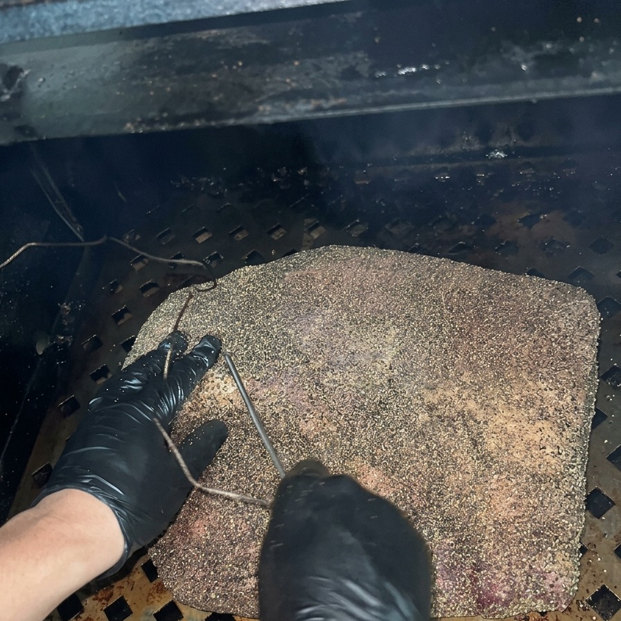 검은 장갑을 낀 사람이 어두운 훈연기 내부에서 양념된 브리스킷의 가장 두꺼운 부위에 금속 온도계 탐침을 꽂는 모습.