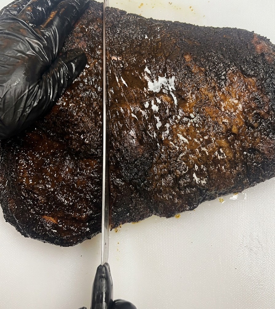 흰색 커팅 보드 위에서 검은 장갑을 낀 손으로 바크가 잘 잡힌 어두운 훈제 브리스킷을 고정하고 긴 칼로 고기를 일정하게 썰어내는 모습.