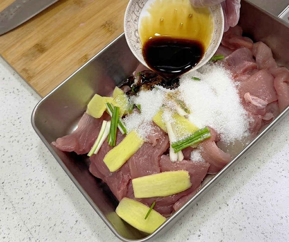 넓은 쟁반에 담긴 얇게 썬 돼지고기 위에 간장, 편 썰어둔 생강, 대파 등 양념을 붓고 있는 모습.