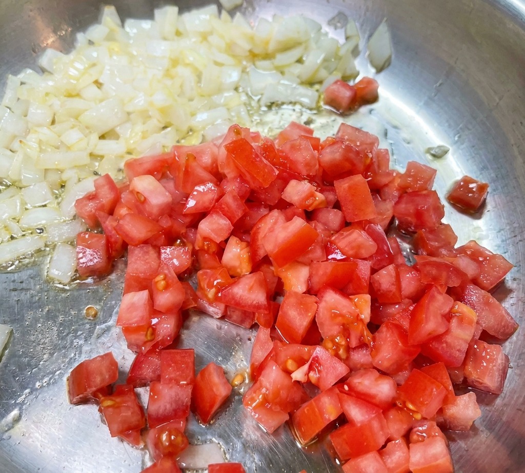 반투명하게 볶아진 양파가 있는 팬에 다진 붉은 토마토를 넣고 섞는 모습.