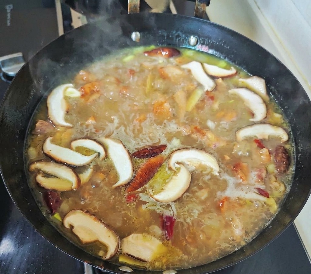 검은 웍 안에서 슬라이스한 표고버섯과 건고추가 황금빛 닭고기 육수와 함께 보글보글 끓고 있는 모습
