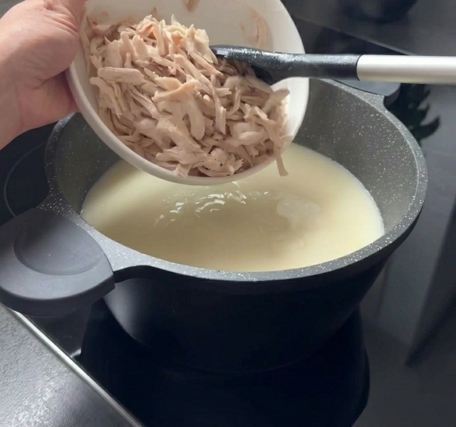 하얀 볼에 담긴 알맞게 찢은 닭고기 살을 크리미한 하얀 수프 냄비에 넣는 모습.