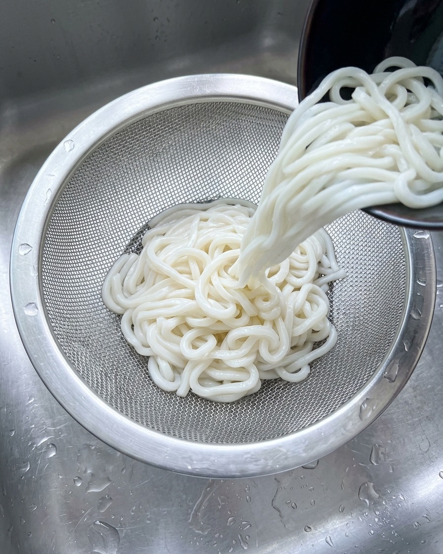 싱크대에서 흐르는 찬물 아래 스테인리스 체에 담긴 흰 우동면을 헹구는 모습