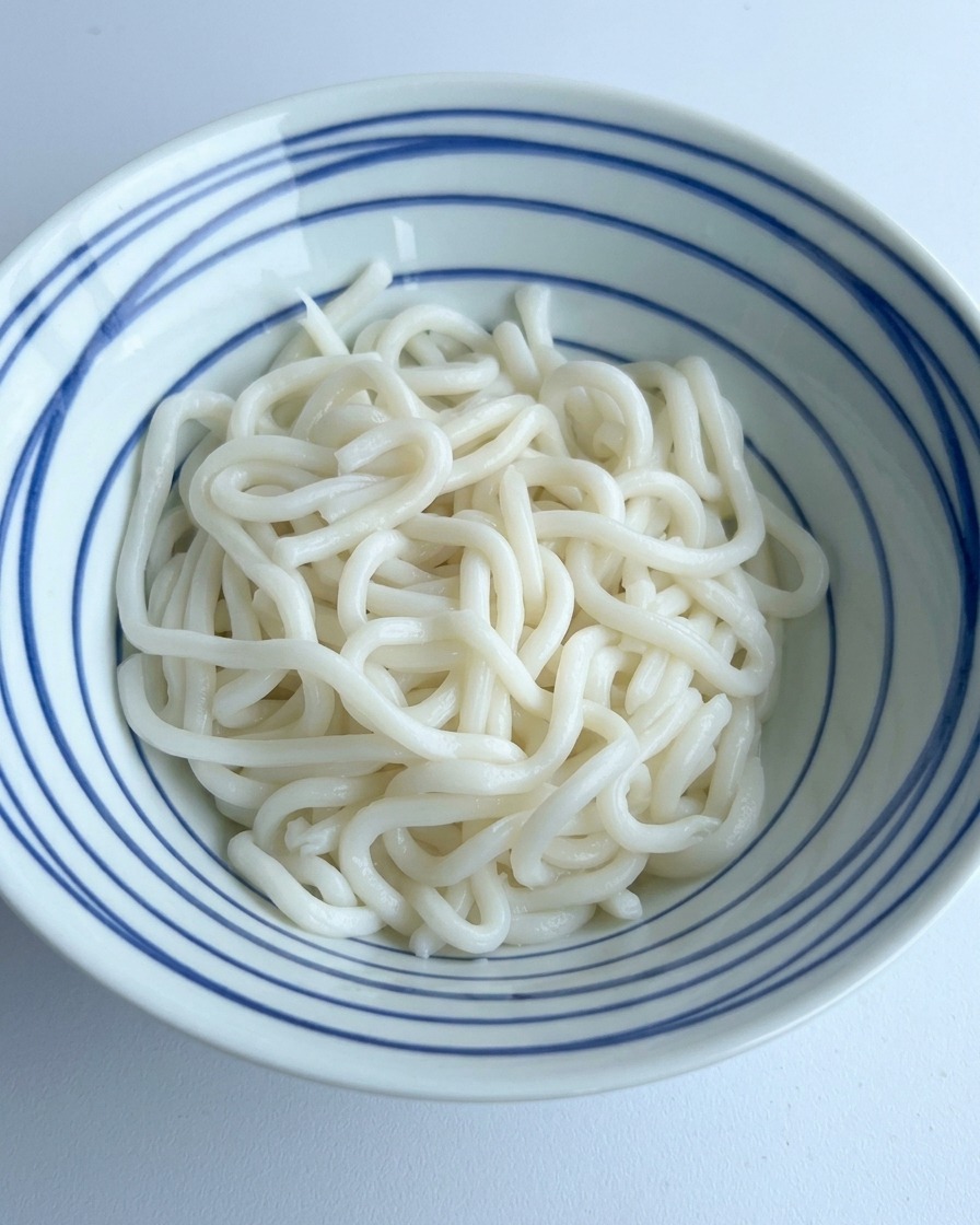 푸른 줄무늬 그릇에 담긴 흰 우동면 위로 진한 간장과 양념을 붓는 모습