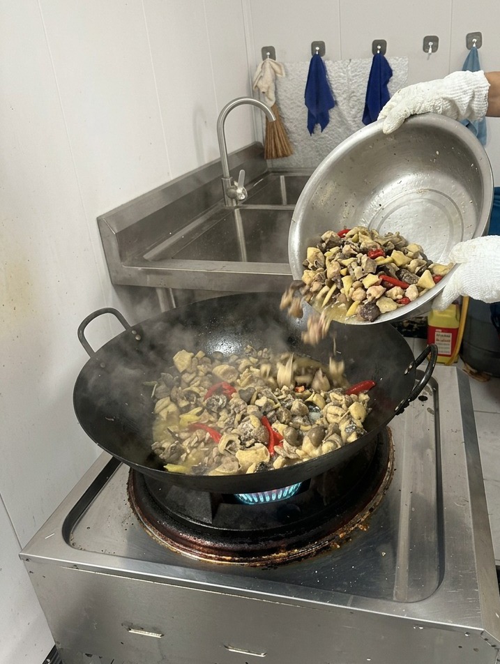 구워진 닭고기와 향신료가 담겨 김이 나고 있는 웍에 조리용 용기로 맑은 끓는 물을 붓고 있는 모습