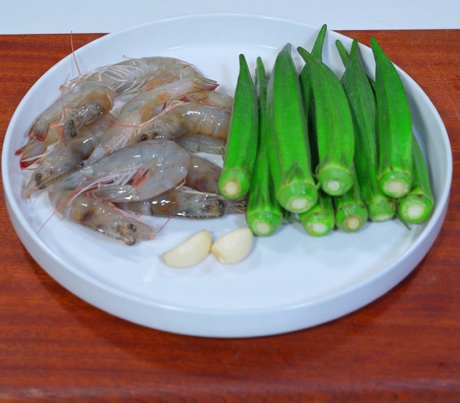 나무 도마 위에 신선한 생새우, 초록색 오크라, 마늘 두 쪽이 흰 접시에 담겨 있는 모습.