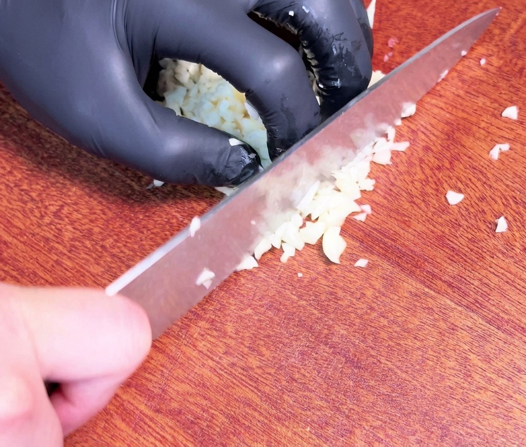 검은색 장갑을 낀 손이 어두운 나무 도마 위에서 날카로운 셰프 나이프로 생마늘을 잘게 다지는 모습.