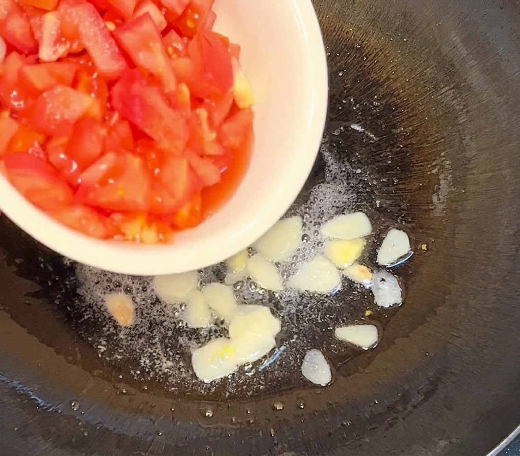 노릇하게 볶아진 마늘 슬라이스가 담긴 팬에 흰 볼에서 다진 토마토를 붓는 모습