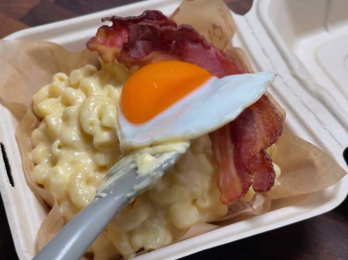 크리미한 맥앤치즈 위에 구운 베이컨을 올리고, 그 위에 주걱으로 계란 프라이를 조심스럽게 얹는 모습.