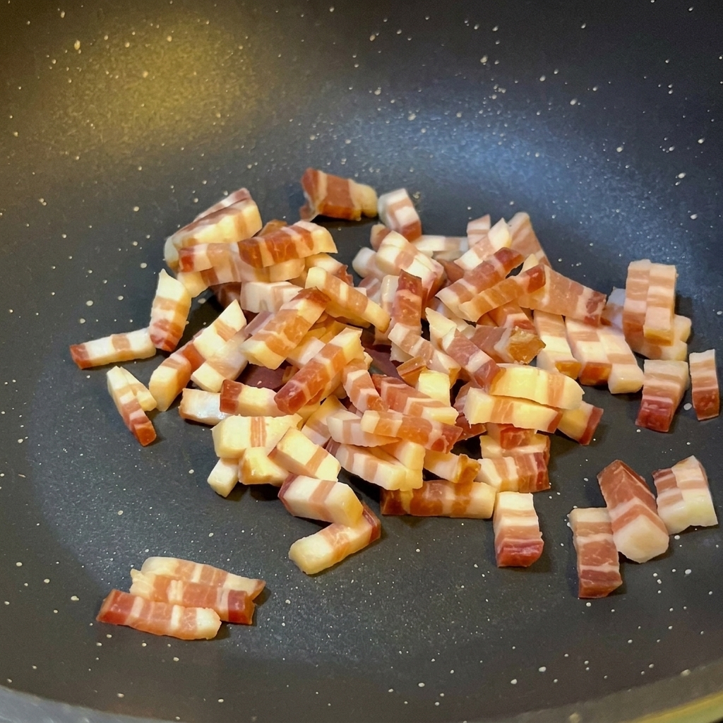 검은색 코팅 팬에서 이탈리아식 판체타 큐브가 기름을 내며 가장자리가 노릇하게 구워지고 있는 모습