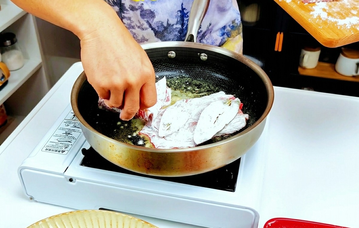 밀가루를 가볍게 묻힌 송아지 고기 조각들을 녹은 버터가 끓는 뜨거운 팬에 넣는 손.
