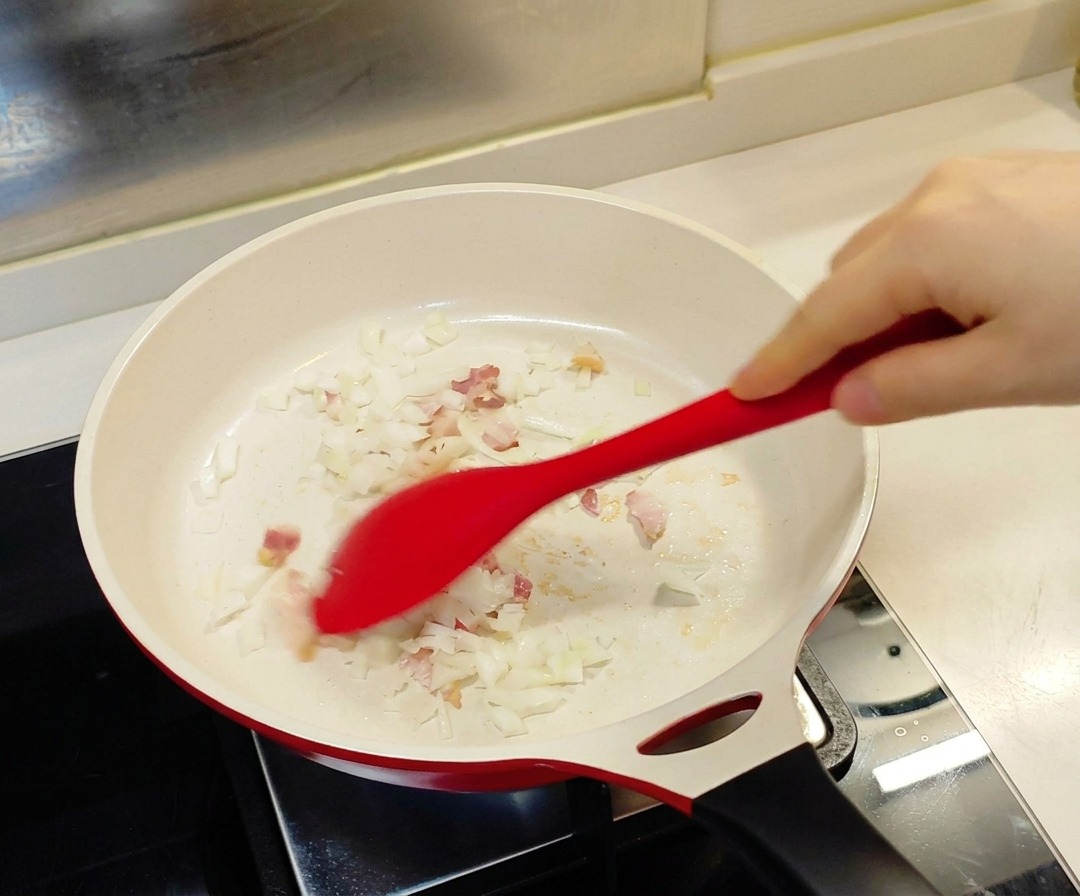 하얀 세라믹 코팅 팬에서 빨간색 실리콘 주걱으로 잘게 썬 양파와 베이컨을 볶는 모습.