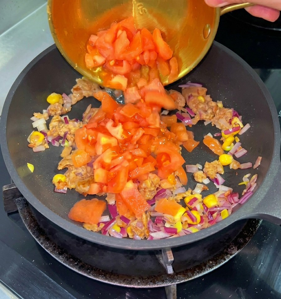 볶은 소고기, 옥수수, 적양파가 담긴 팬에 작은 놋그릇에 담긴 다진 붉은 토마토를 붓는 모습