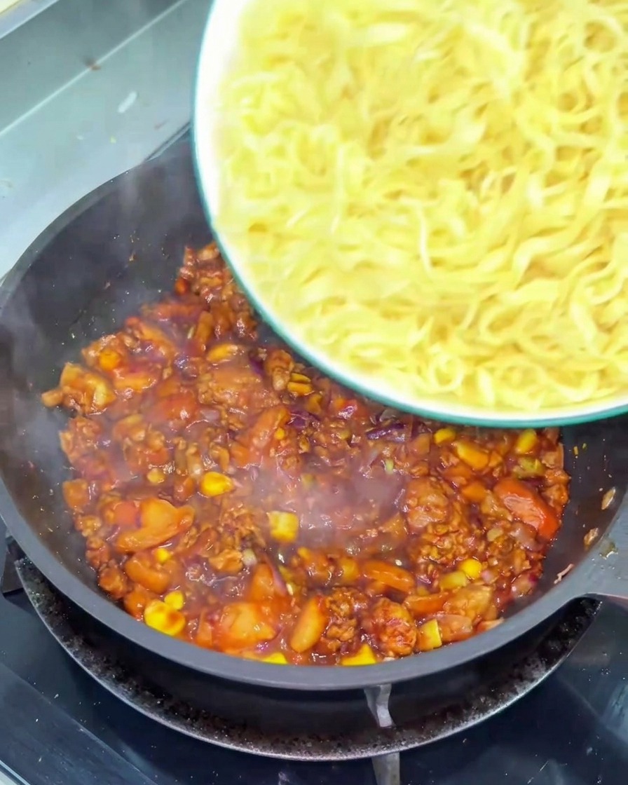 걸쭉하고 붉은 토마토 소고기 소스가 가득한 프라이팬에 물기를 뺀 꼬들꼬들한 노란 밀면을 넣는 모습