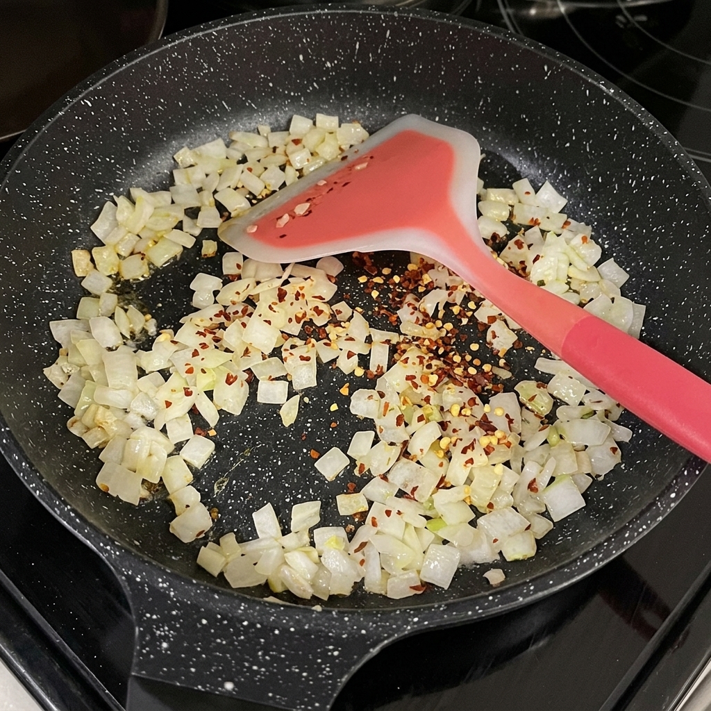 검은색 프라이팬 속 투명하게 볶아진 양파 위에 말린 빨간 고춧가루가 뿌려져 있는 모습.
