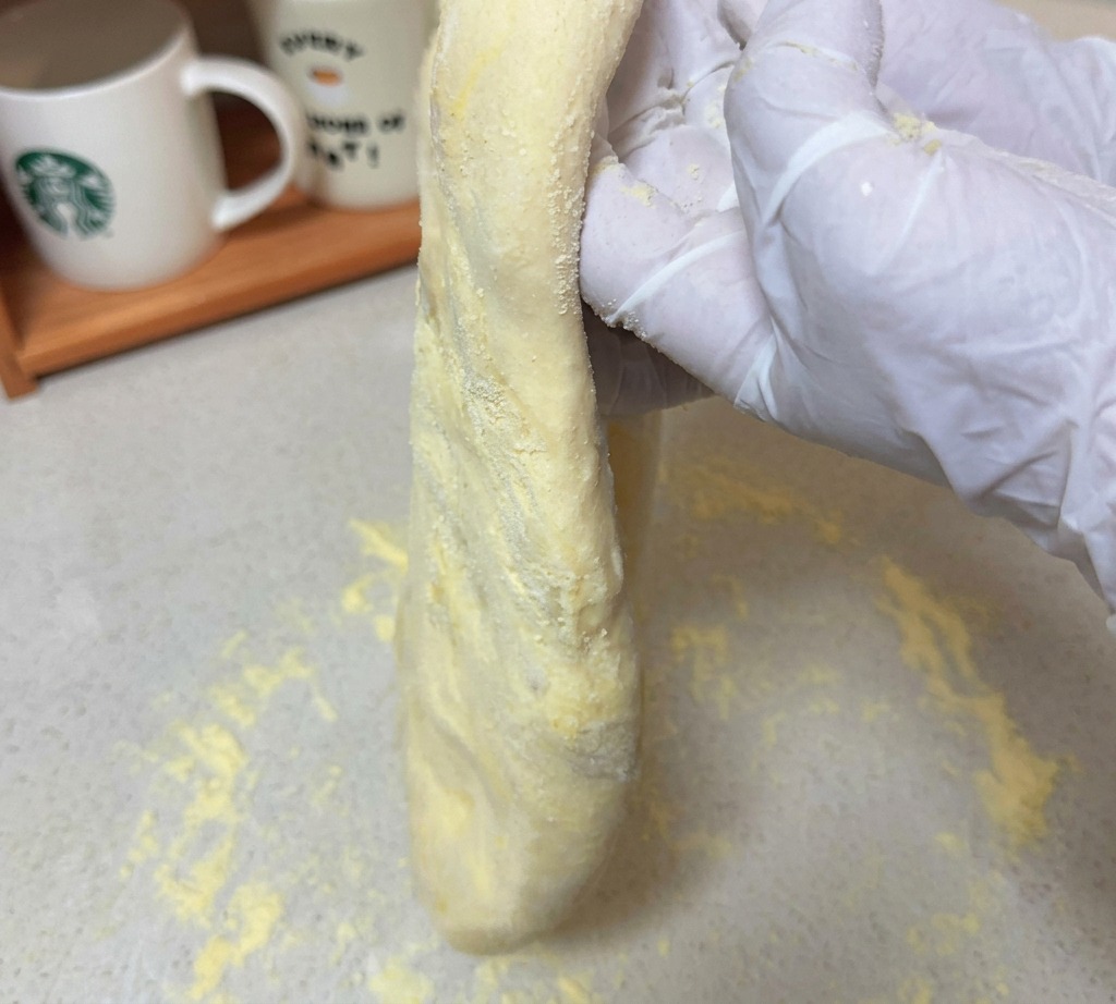 밀가루가 뿌려진 조리대 위로 장갑을 낀 손이 길게 늘려진 생피자 도우를 들어 올리고 있는 모습