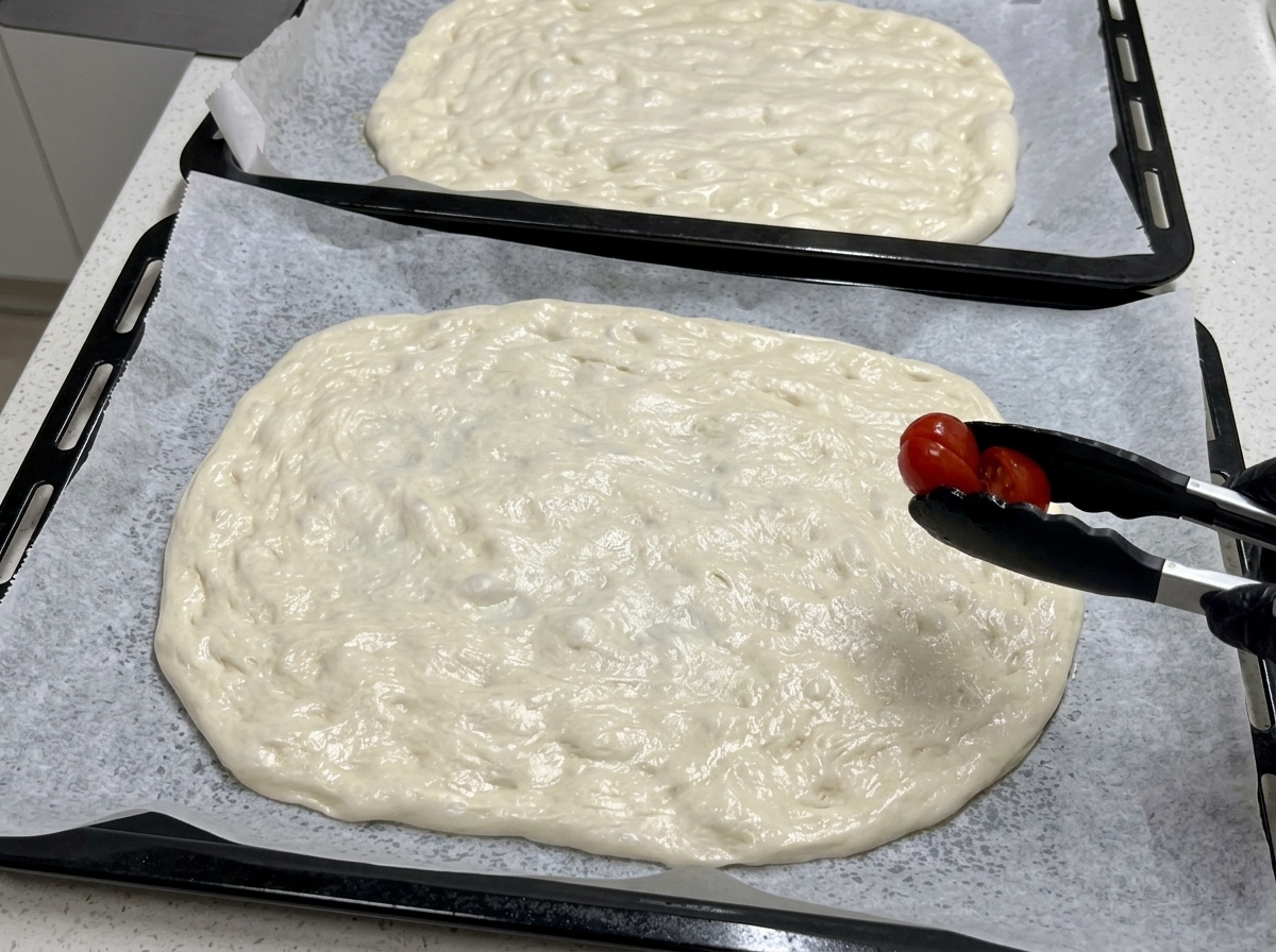 검은색 요리용 집게로 반으로 자른 방울토마토를 넓게 편 피자 생지 위에 올리는 모습.