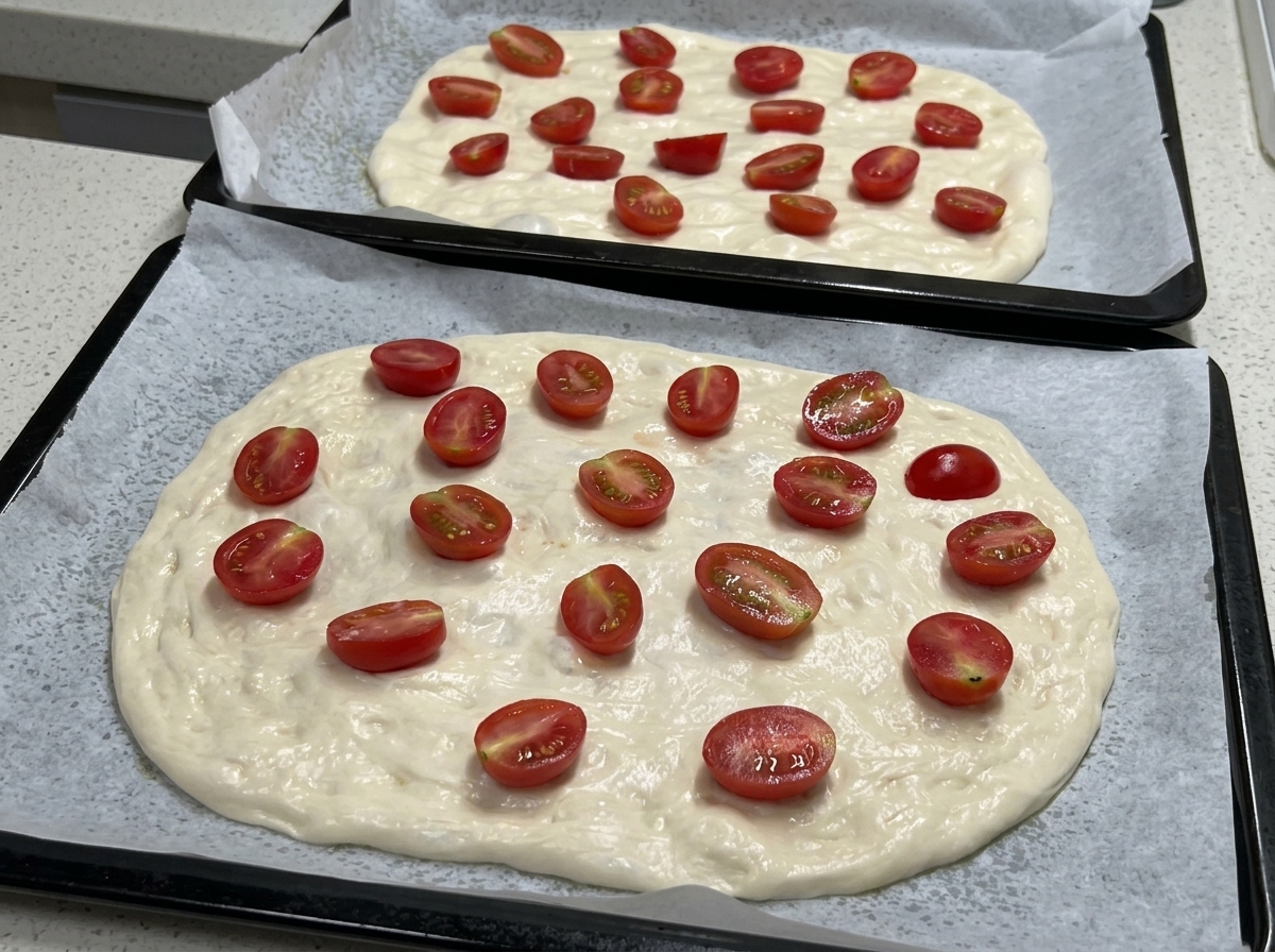 반으로 자른 방울토마토가 가득 올라간 채 시즈닝을 기다리는 피자 반죽이 담긴 두 개의 베이킹 트레이.