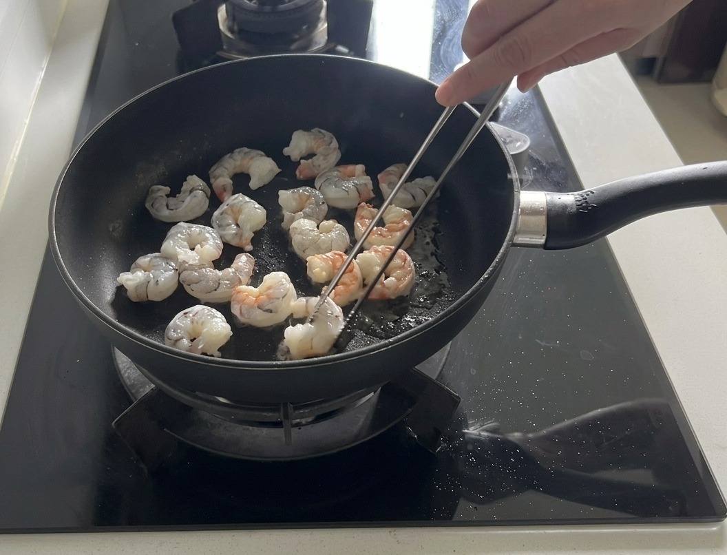 올리브 오일을 두른 어두운 프라이팬에서 새우가 붉고 불투명하게 익어가고 있는 모습.