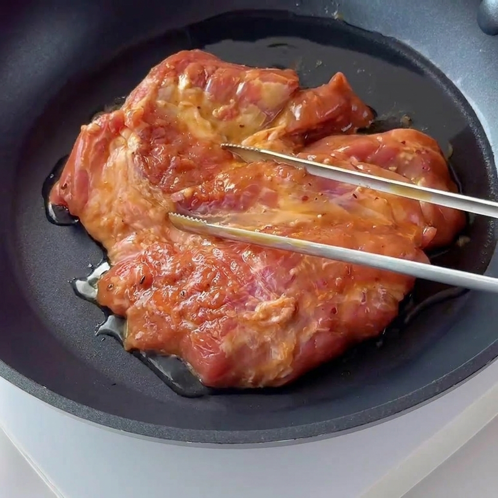 짙은 색의 코팅 프라이팬에서 양념된 돼지고기가 지글지글 노릇하게 구워지는 모습.