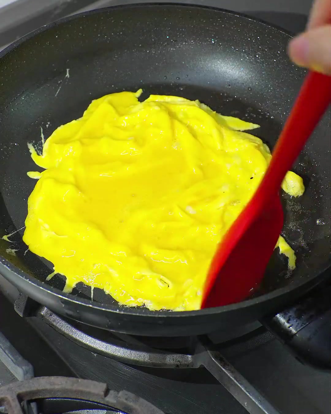 검은색 프라이팬에서 빨간색 주걱으로 반쯤 익은 밝은 노란색의 스크램블 에그를 접는 모습.