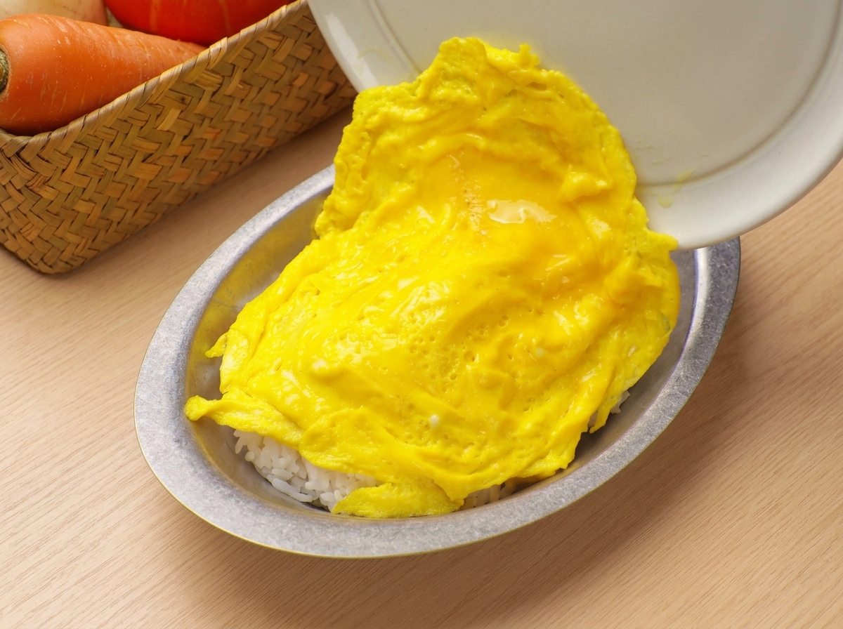 부드럽고 푹신한 스크램블 에그를 프라이팬에서 흰 밥이 담긴 그릇으로 조심스럽게 미끄러지듯 옮기는 모습.