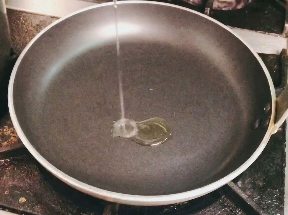 가스레인지 위의 빈 검은색 프라이팬에 식용유를 가늘게 붓는 모습.