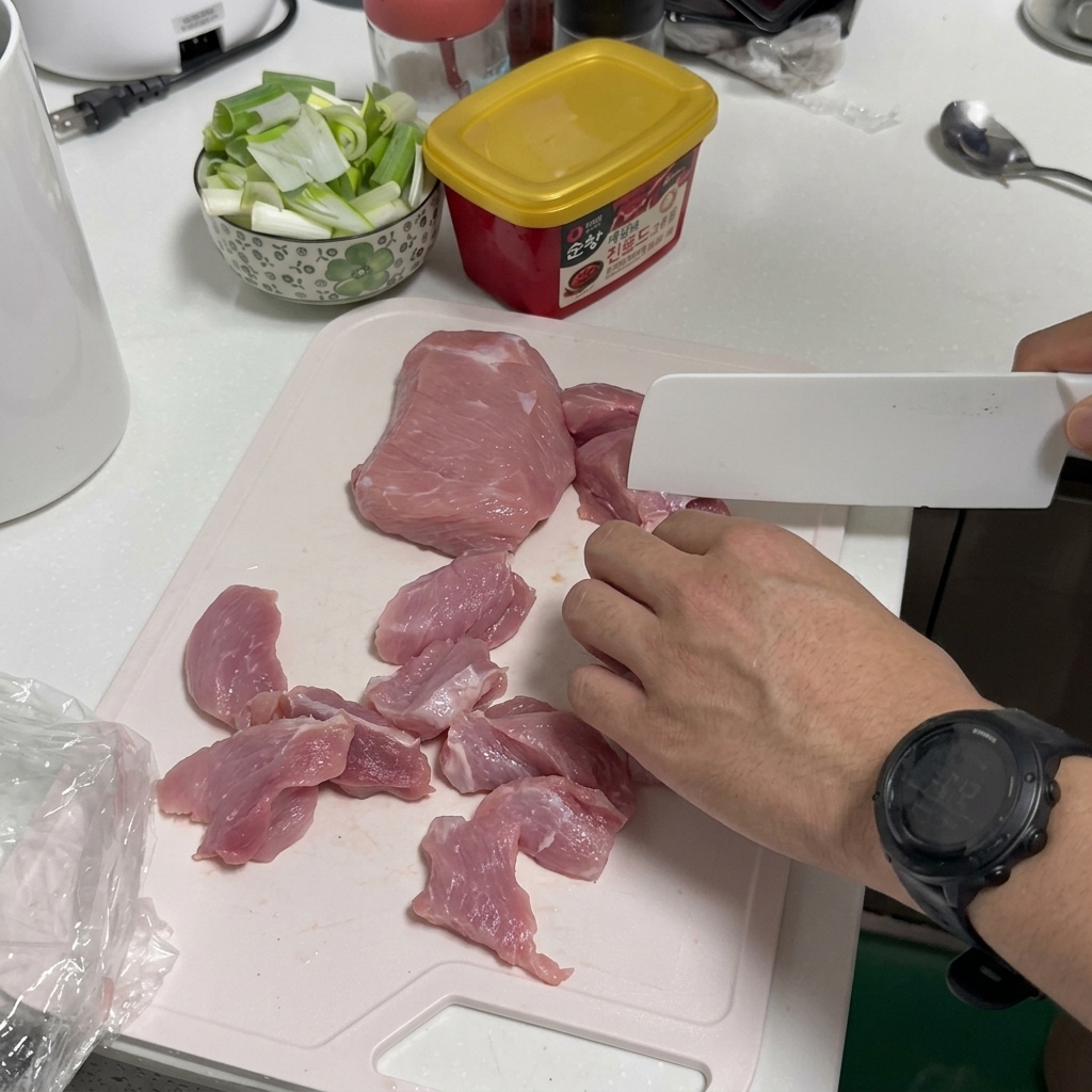 흰 도마 위에서 흰색 식칼로 생 돼지고기를 얇게 써는 모습.