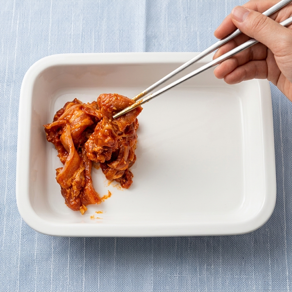양념한 삼겹살을 쇠젓가락을 이용해 흰색 오븐 용기에 담는 모습.