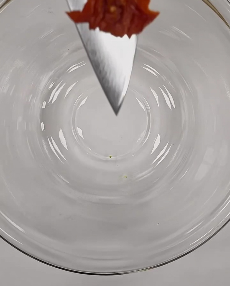 유리 볼 위에서 셰프 나이프 끝에 올려진 잘게 썬 붉은 썬드라이 토마토 한 조각