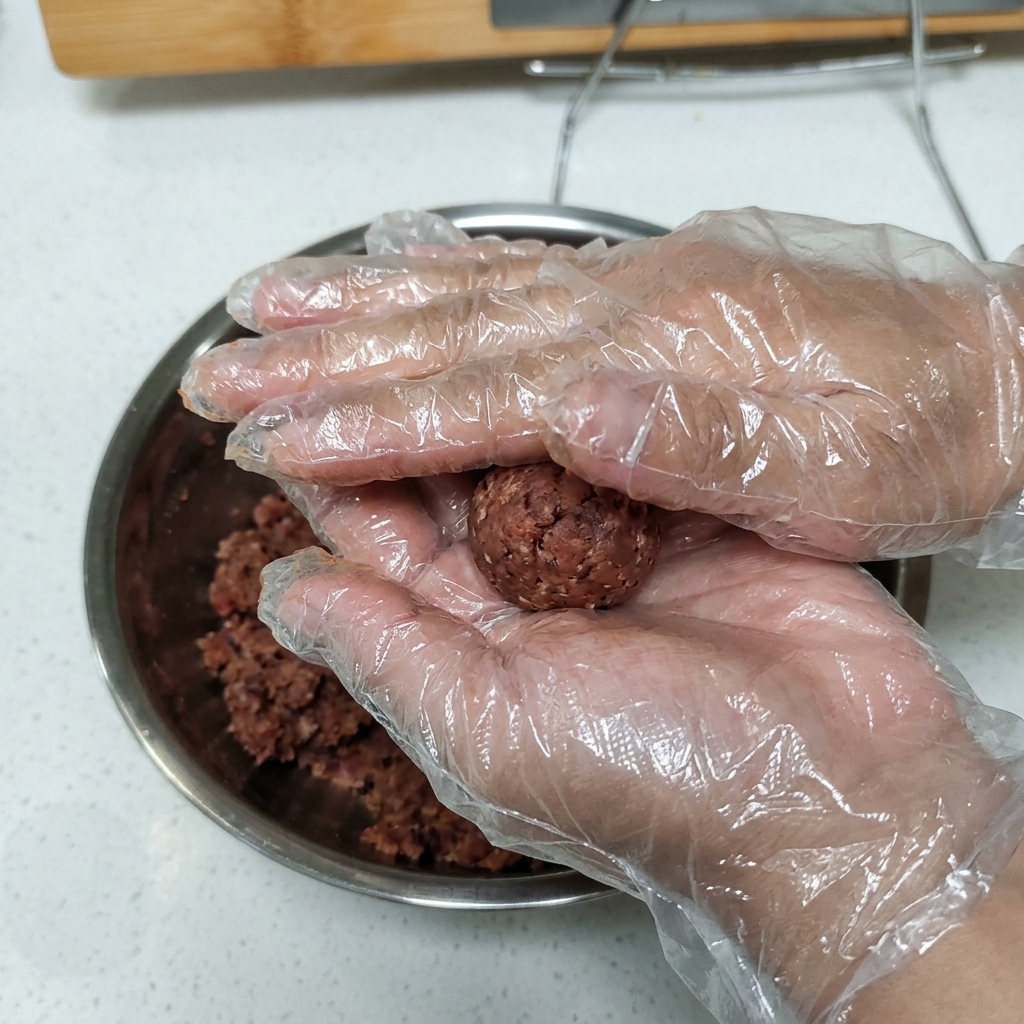장갑을 낀 손으로 양념된 다진 고기 반죽을 동그란 패티 모양으로 누르는 모습.