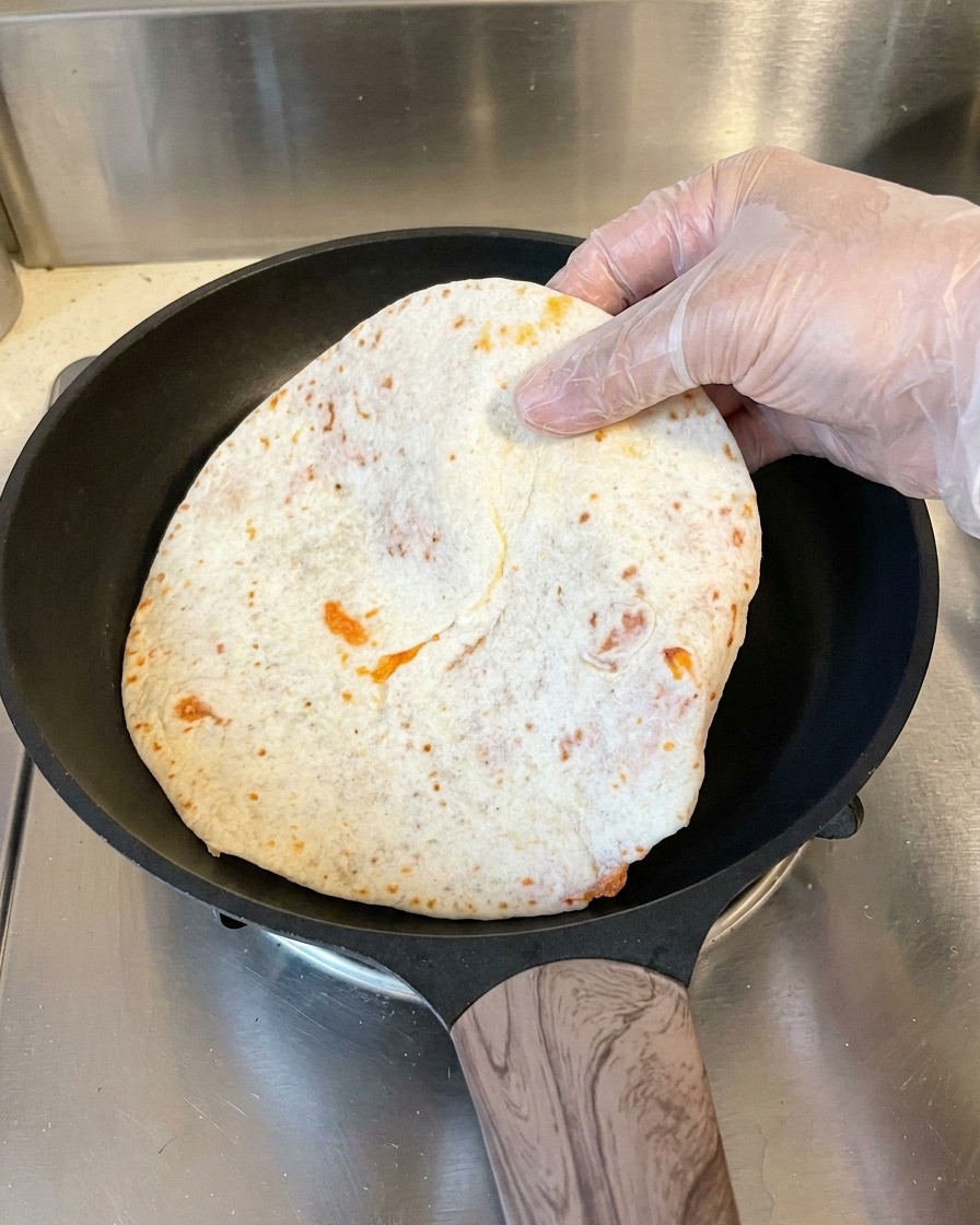 비닐장갑을 낀 손이 납작하게 만들어 종이 호일에서 분리한 파전을 뜨겁게 달궈진 검은색 코팅 팬에 올리는 모습.