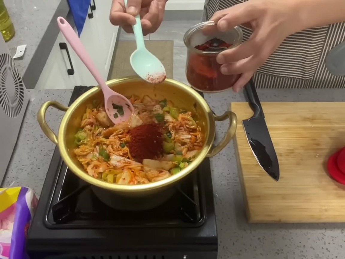 볶은 김치 혼합물에 선명한 붉은 고춧가루 한 숟가락을 넣는 모습.