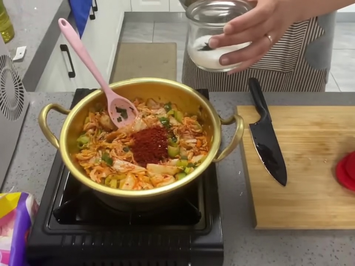 냄비에서 양념 된 김치 혼합물에 하얀 설탕 반 숟가락을 넣는 모습.