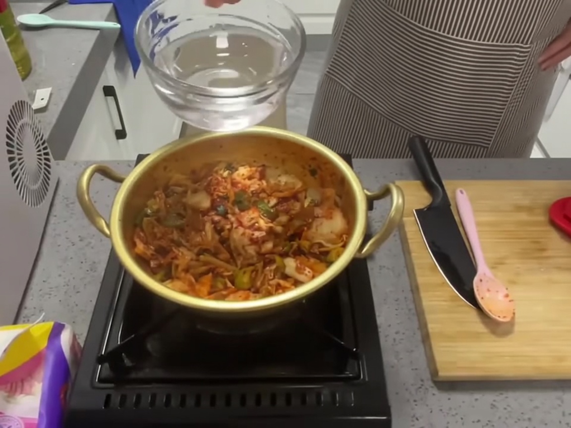 유리 계량컵에 담긴 맑은 물을 볶은 김치와 향신채가 있는 냄비에 붓는 모습.