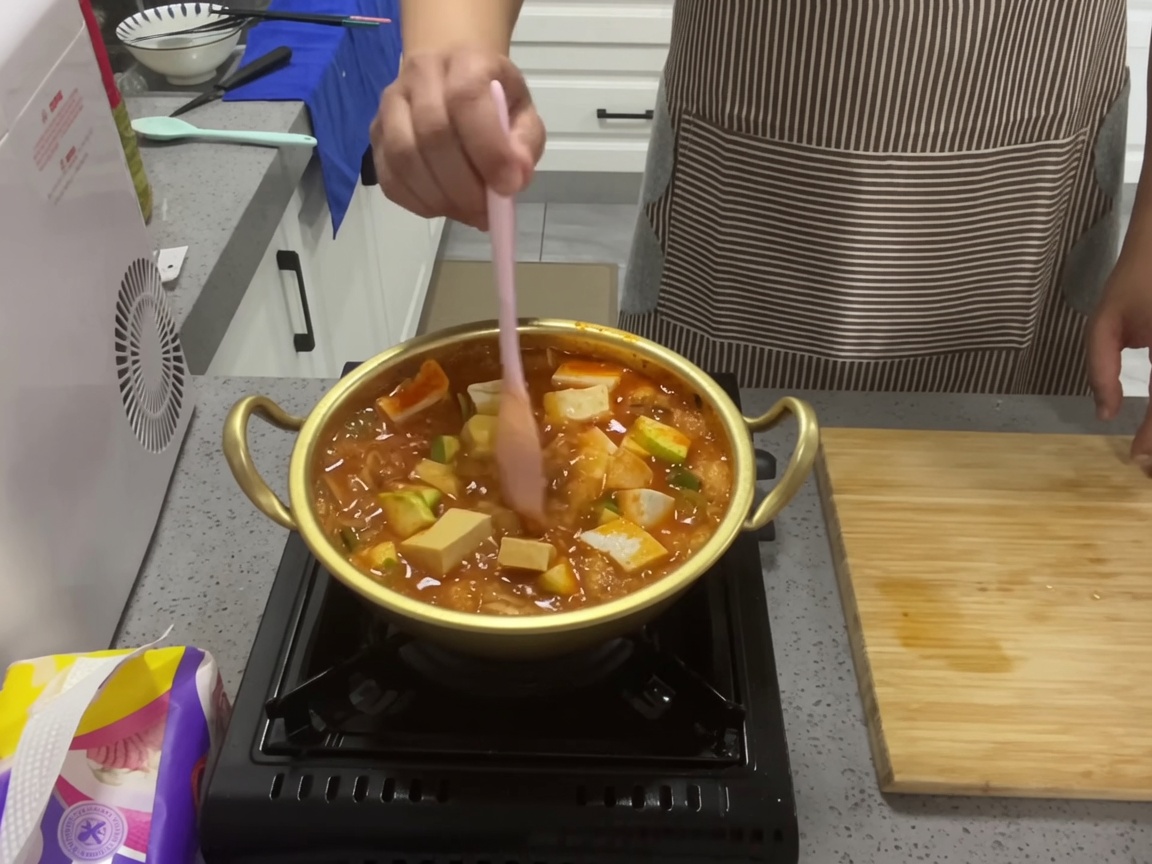 양은냄비에서 보글보글 끓고 있는 참치 김치찌개를 분홍색 실리콘 숟가락으로 저으며 두부와 애호박을 섞는 클로즈업 사진.