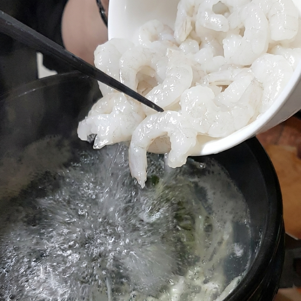 보글보글 끓는 뚝배기 육수에 흰 그릇에 담긴 생새우를 넣는 모습.