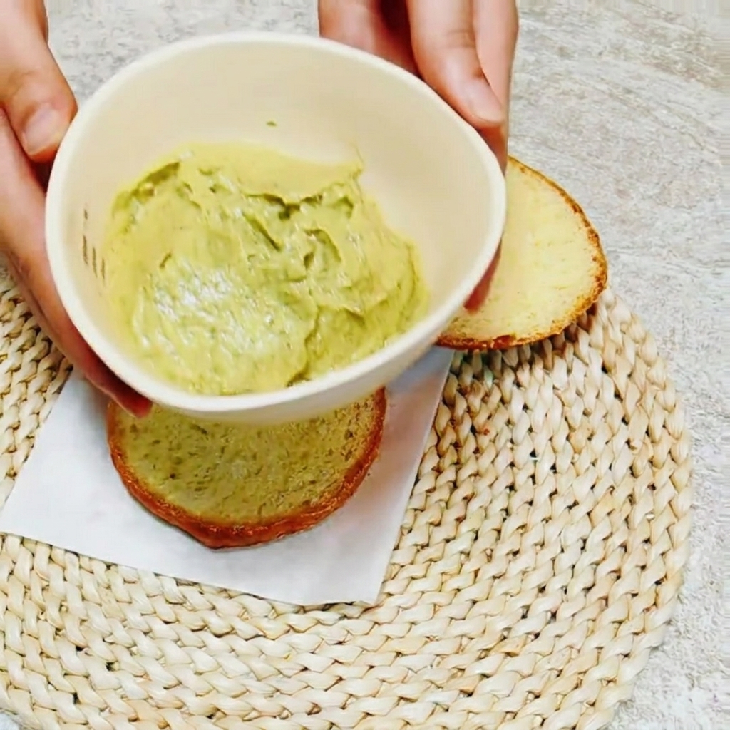 버거 번 단면에 밝은 녹색의 아보카도 바질 소스를 칼로 고르게 펴 바르는 모습.