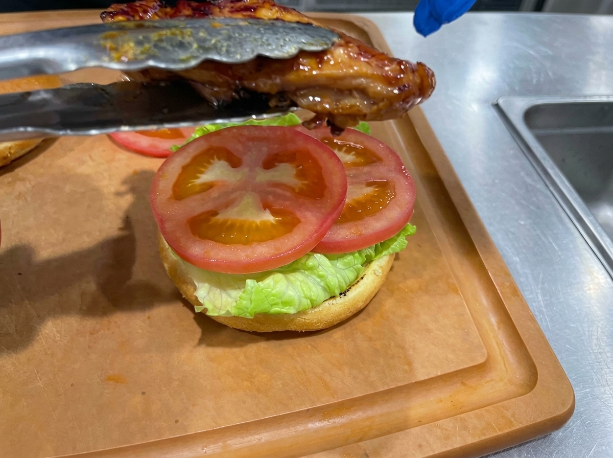 양상추와 토마토가 겹겹이 쌓인 버거 번 위로 구워진 데리야끼 치킨을 집게로 올리는 모습.