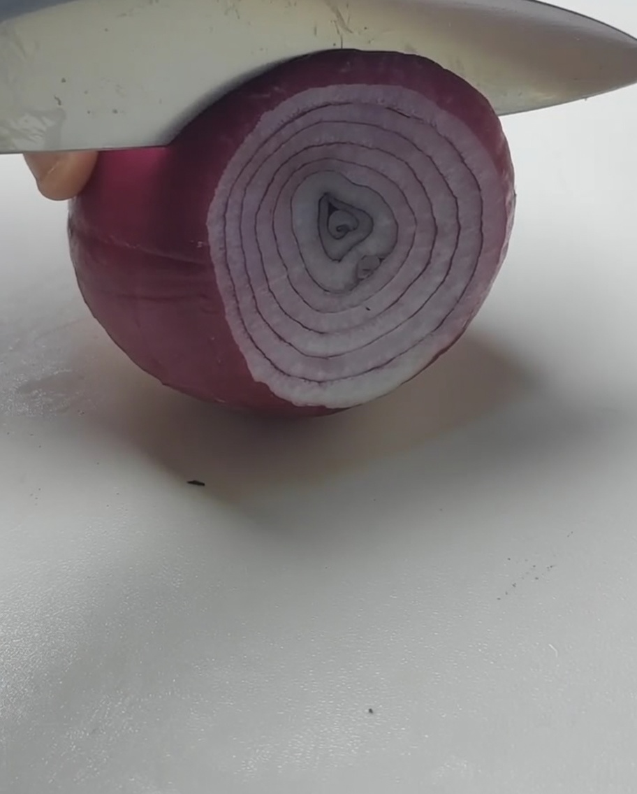 흰 도마 위에서 칼로 적양파를 두툼한 고리 모양으로 써는 모습.