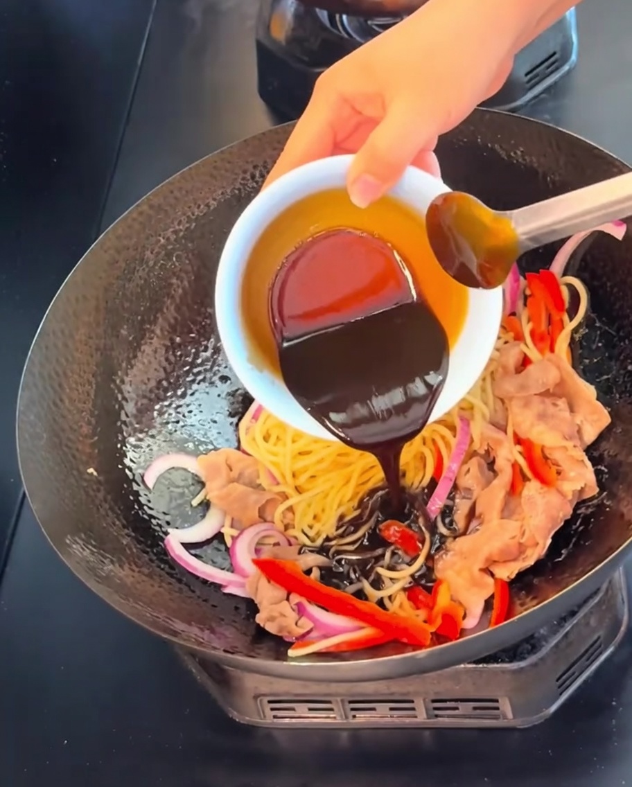진한 블랙페퍼 소스를 웍 안의 면과 재료 위로 붓는 모습.
