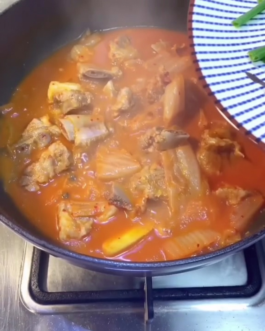 끓고 있는 붉은 김치찌개 위로 신선한 대파, 고수, 얇게 썬 청양고추를 뿌리는 클로즈업 사진.