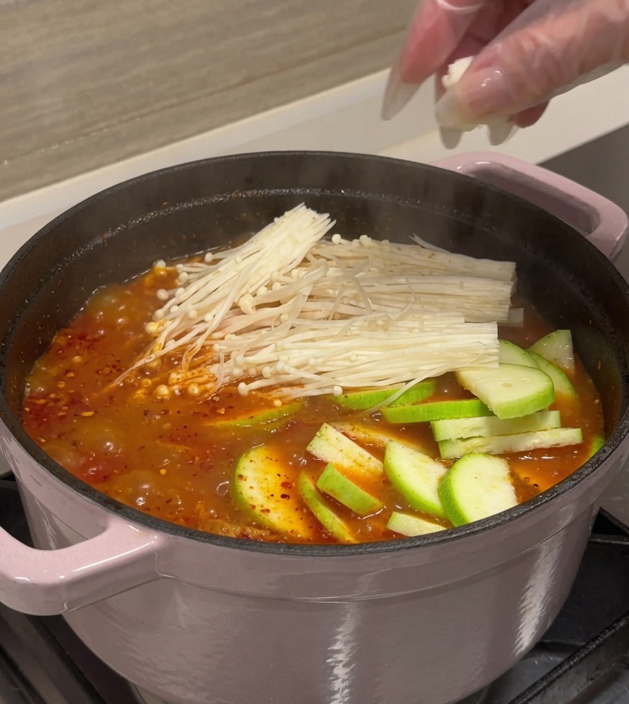 붉게 끓고 있는 김치찌개 냄비에 애호박, 두부, 팽이버섯을 넣는 모습.