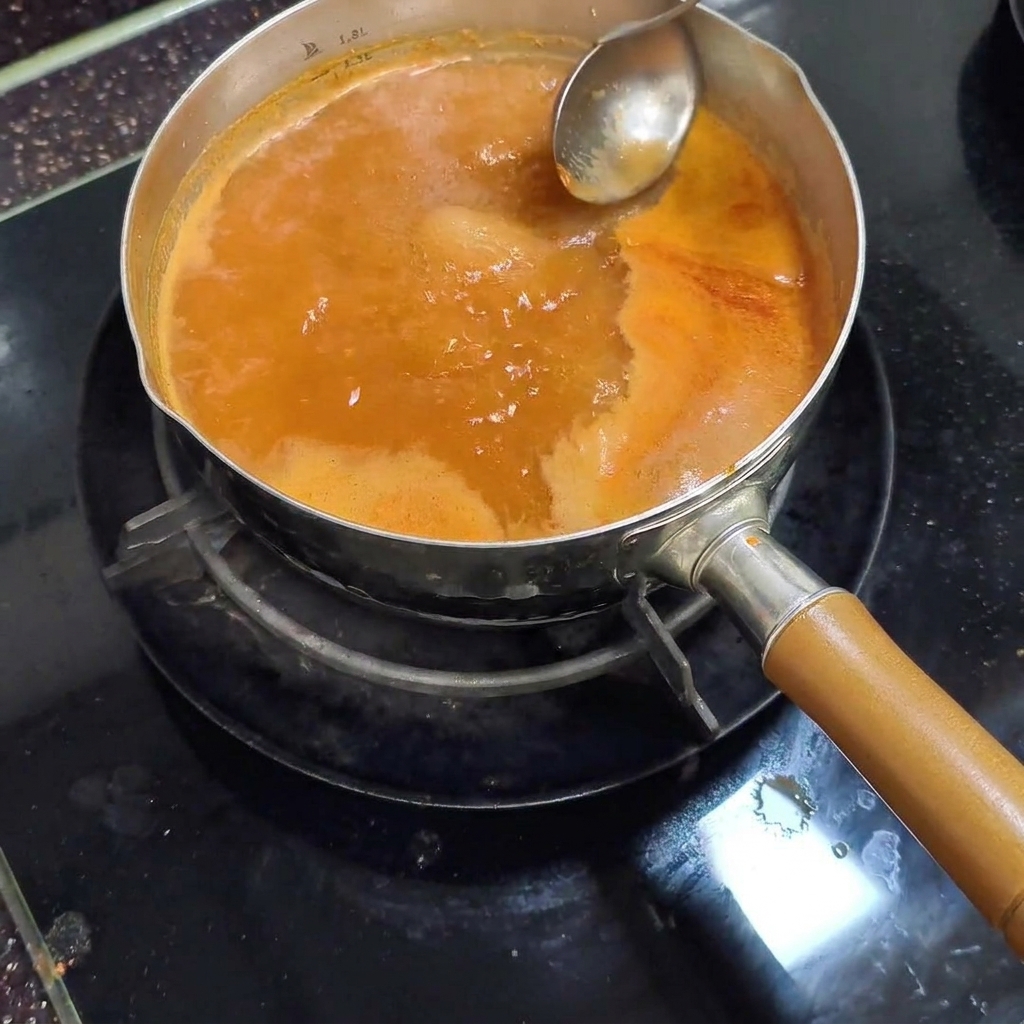 금속 국자로 끓고 있는 붉은 국물 표면의 밝은 색 거품을 걷어내는 모습.